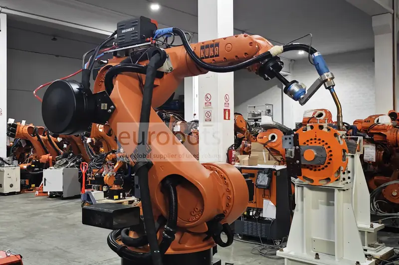 Orange KUKA welding robot holding a welding torch beside a positioner in the Eurobots workshop.