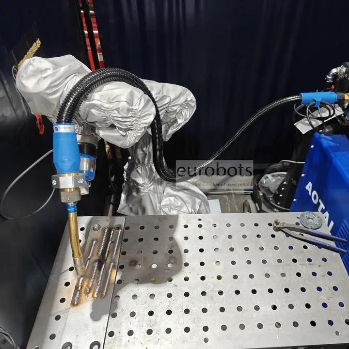 A DOBOT CR10 cobot arm in a protective cover guides a MIG/MAG welding torch over weld seams on a perforated welding table.